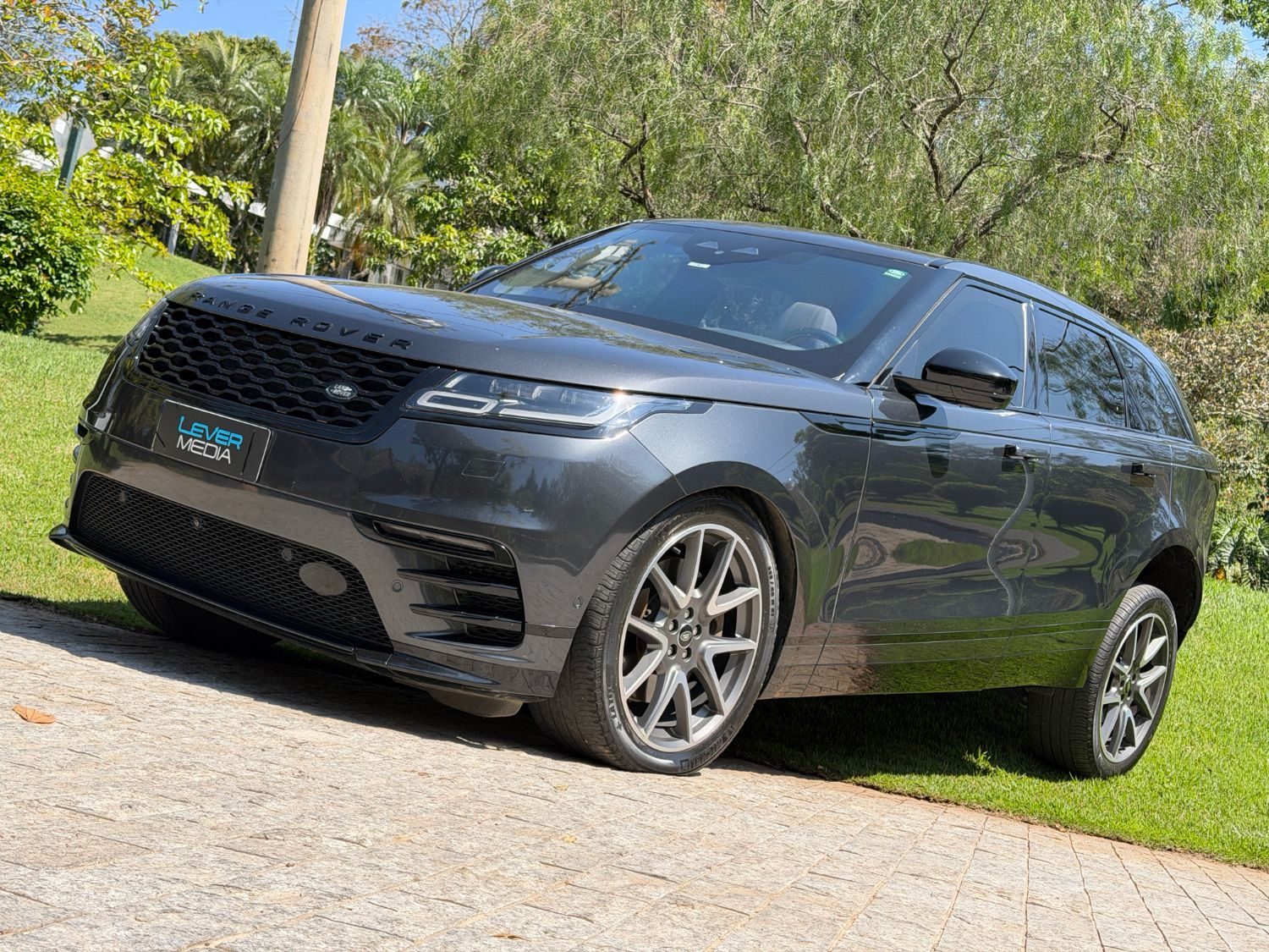 Range Rover Velar cinza do Barone, com a placa Lever Media, estacionado num caminho de pedra cercado de árvores
